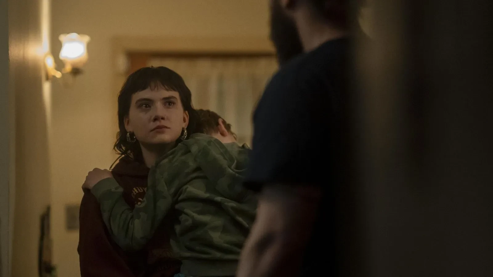 Person standing in a dimly lit hallway holding a small child wearing a camouflage top, while another person stands in the foreground facing them.