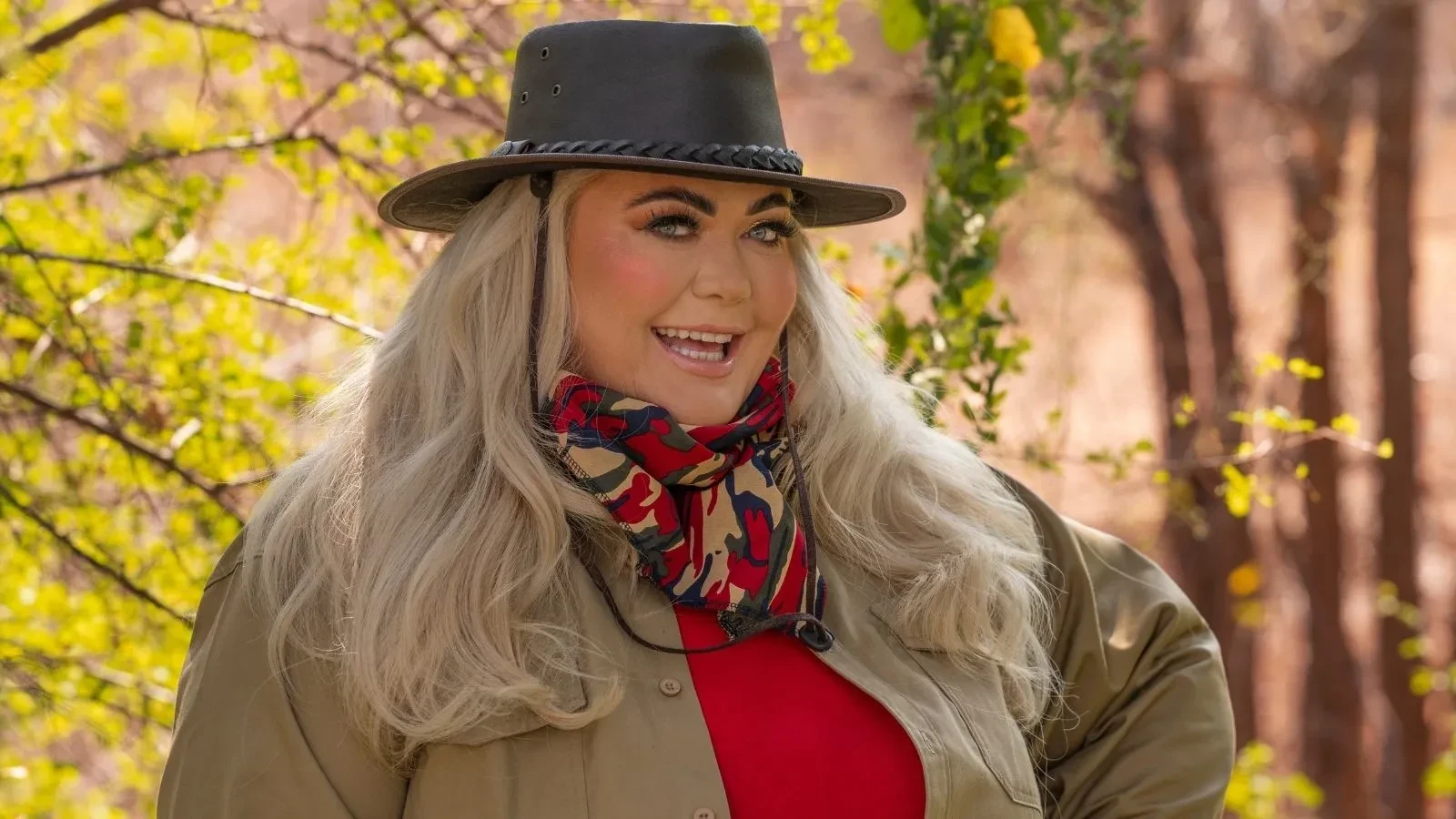 Gemma Collins standing among leafy branches in the South African bush, wearing a red top, khaki shirt, and patterned neck scarf with a wide‑brimmed hat.