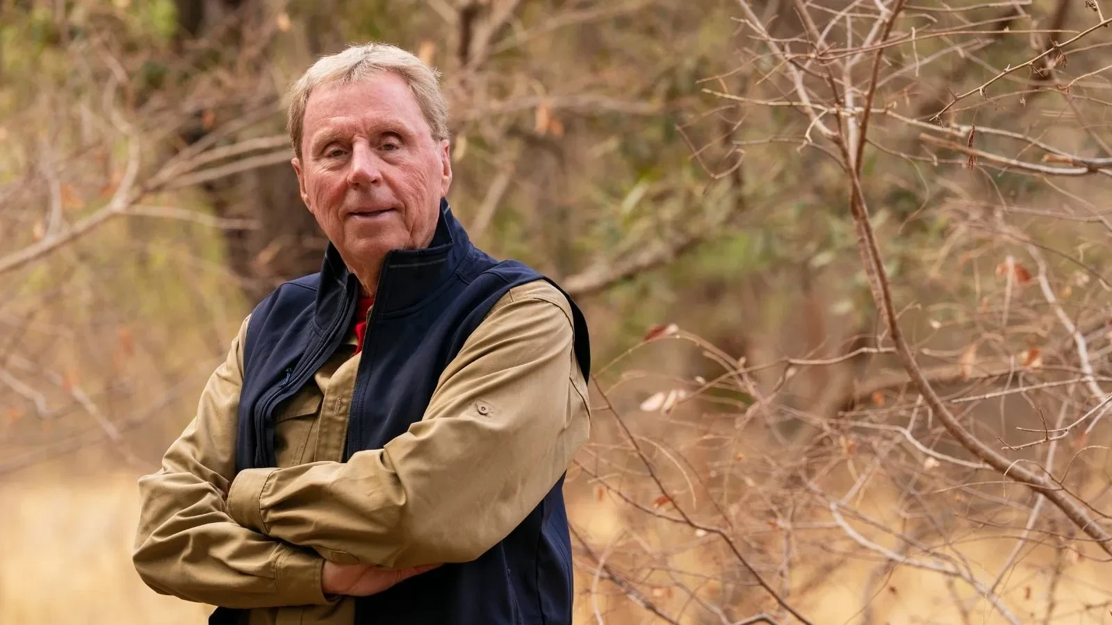 Harry Redknap standing with arms open in a dry woodland area, wearing a red T‑shirt, khaki shirt, and dark vest.