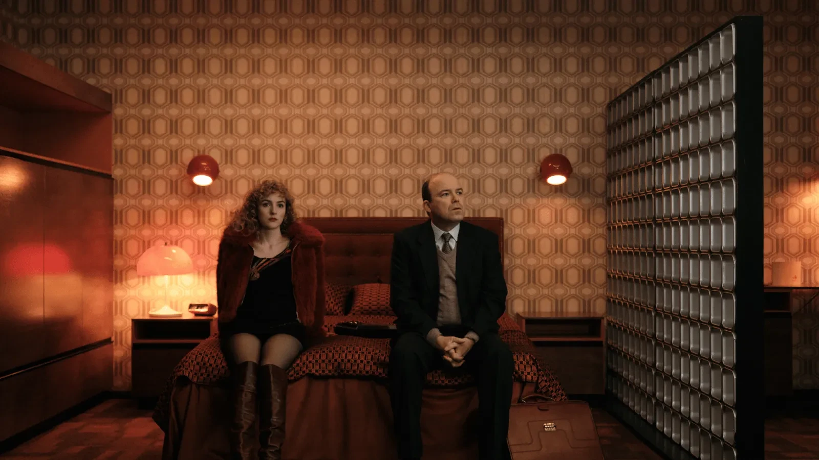 Two people sitting on a bed in a dimly lit vintage-style room with patterned wallpaper, red lamps, and retro décor