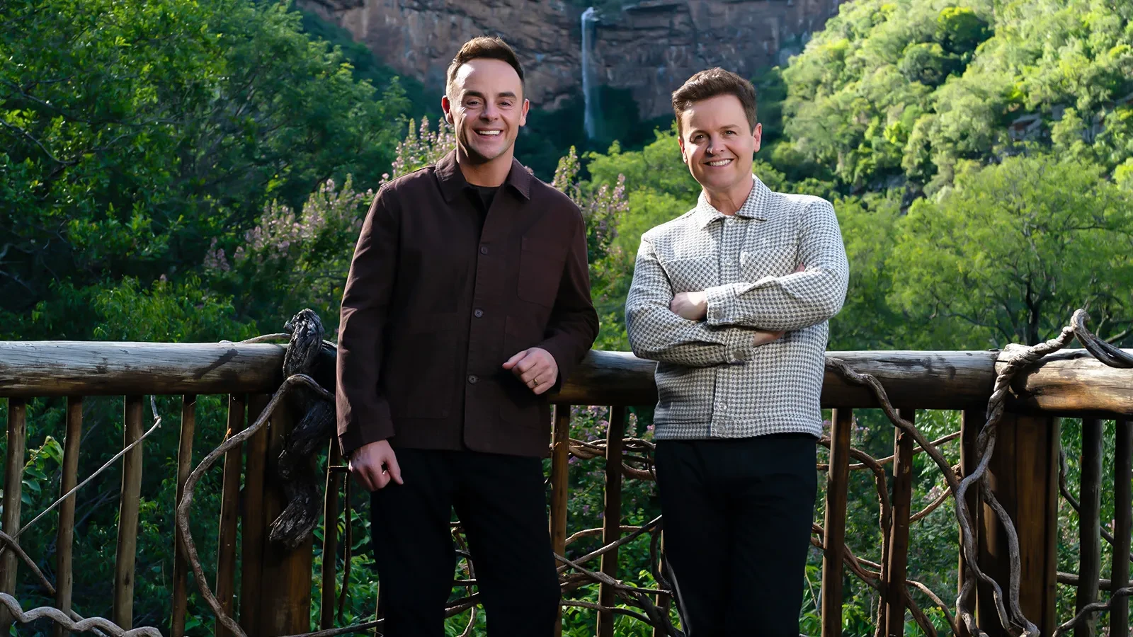 Two people standing on a wooden viewing deck surrounded by lush green forest, with a rocky cliff and waterfall in the background.