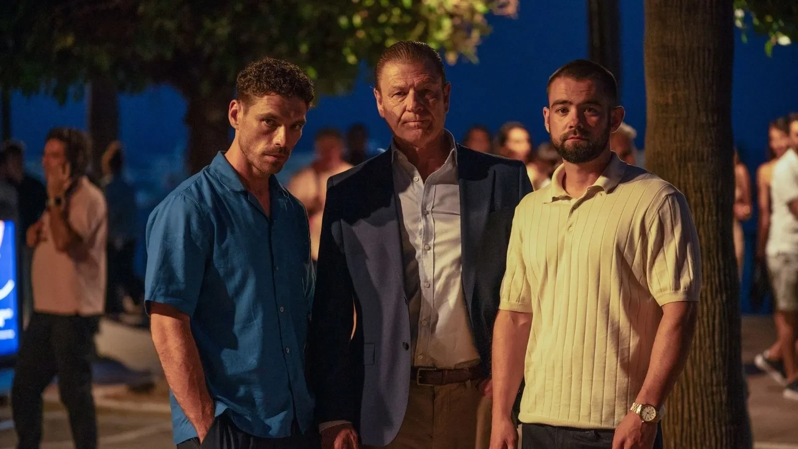 Three people standing together on a lively promenade at night beneath streetlights and trees.