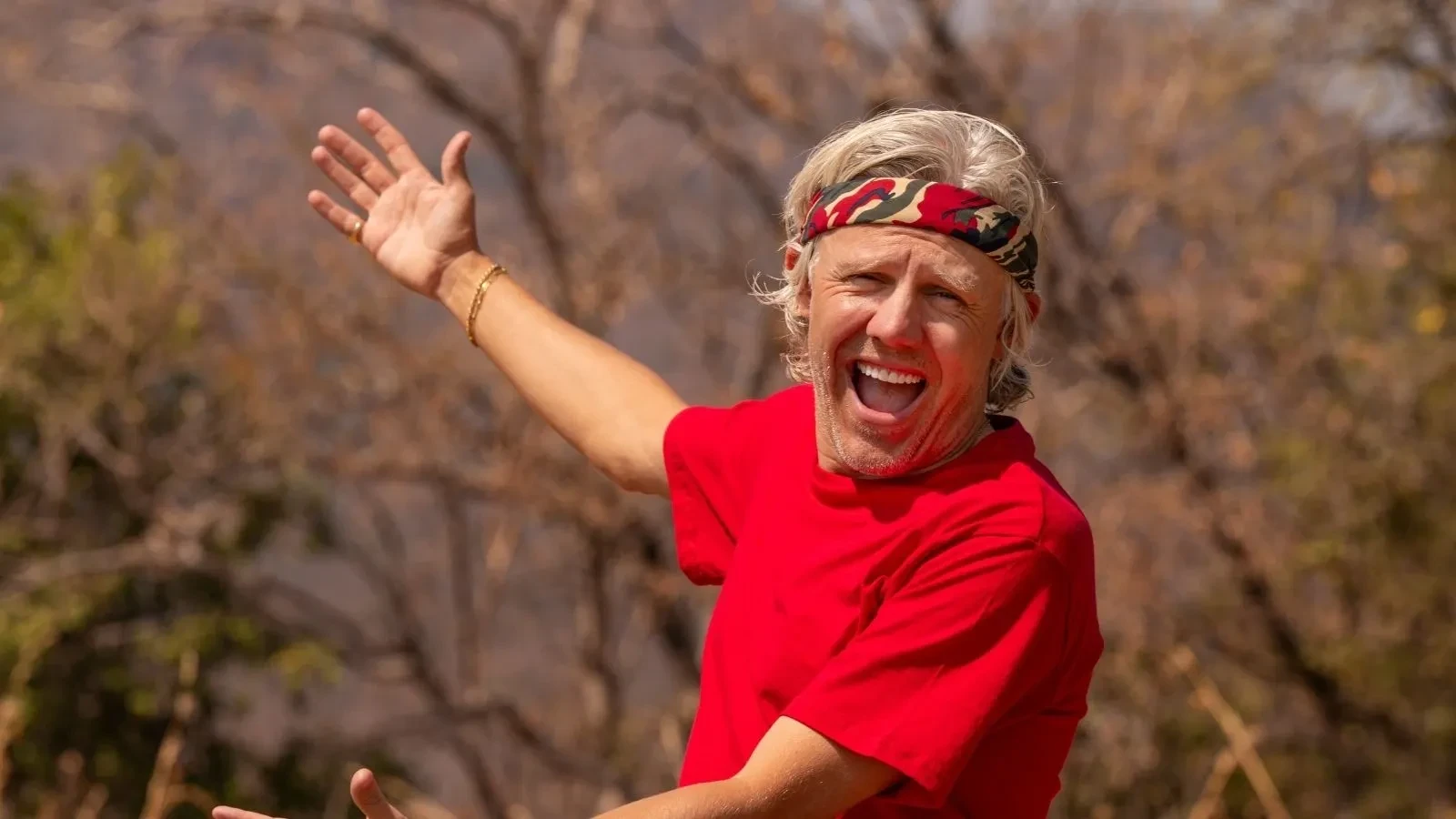 Jimmy Bullard gesturing outward with both arms in a dry, open landscape, wearing a red T‑shirt and patterned bandana.