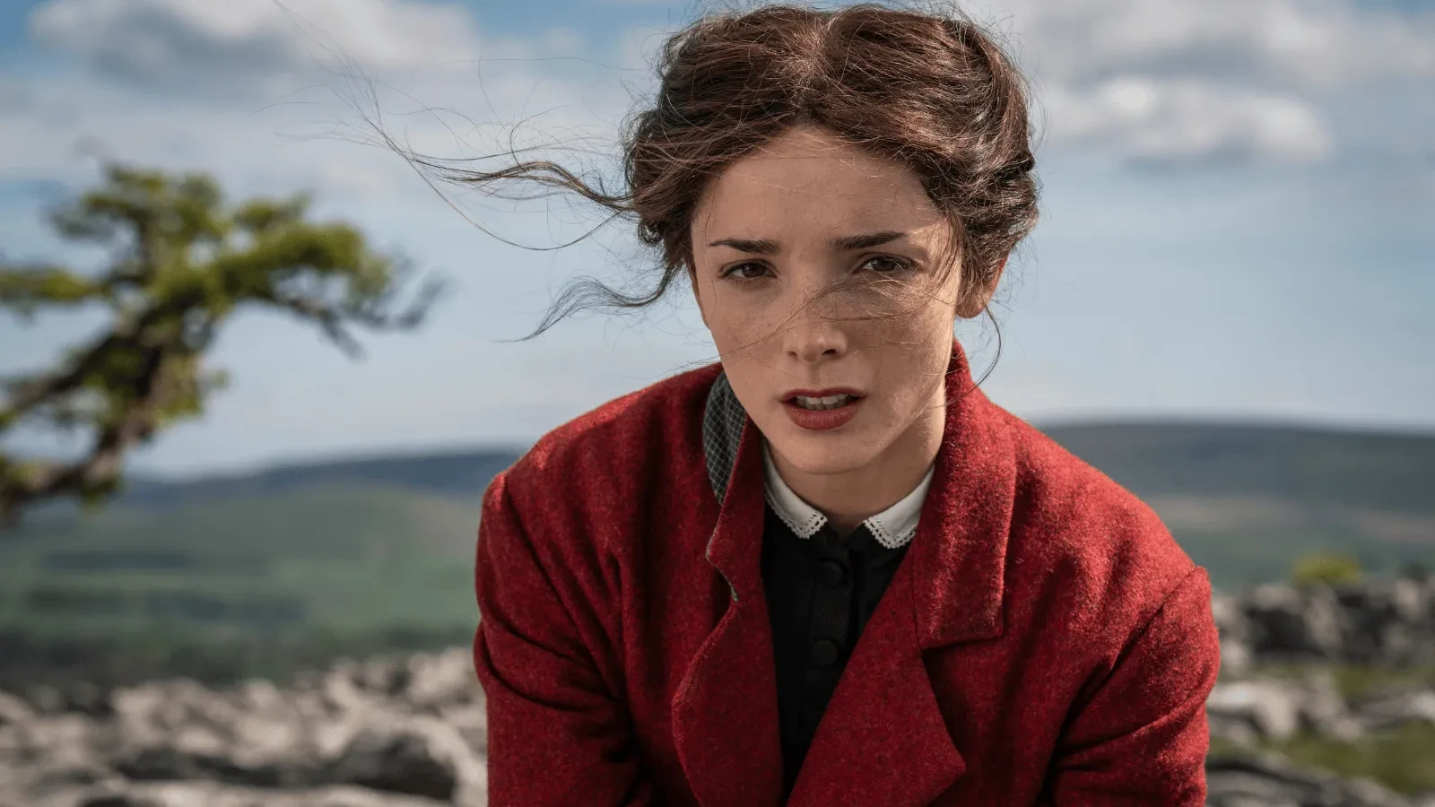 A person wearing a red coat sits outdoors on a rocky landscape with rolling hills and a partly cloudy sky in the background. Loose strands of hair blow in the wind. 