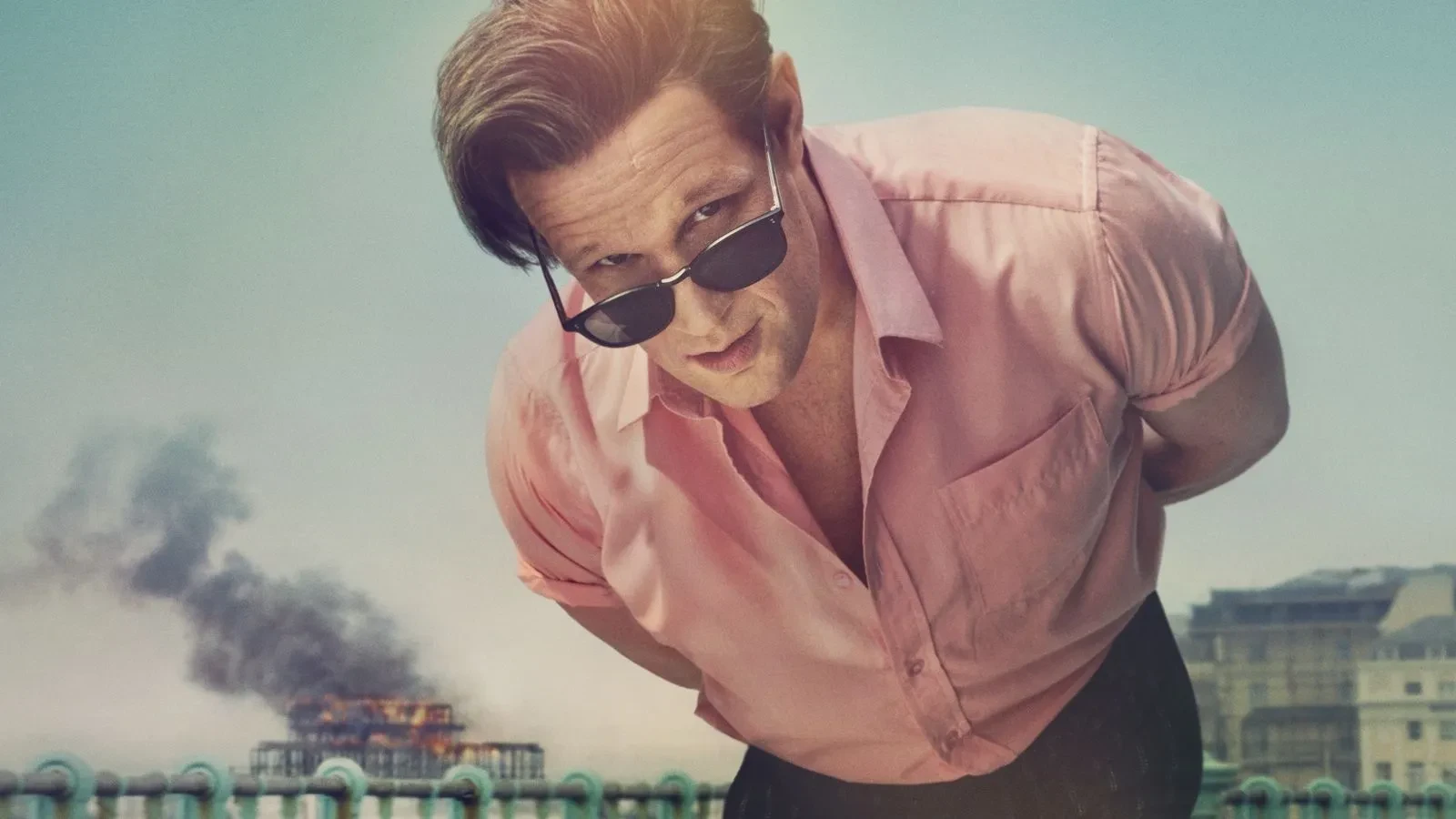 Individual in a pink shirt leaning forward on a seafront promenade with smoke rising from a burning pier in the distance.