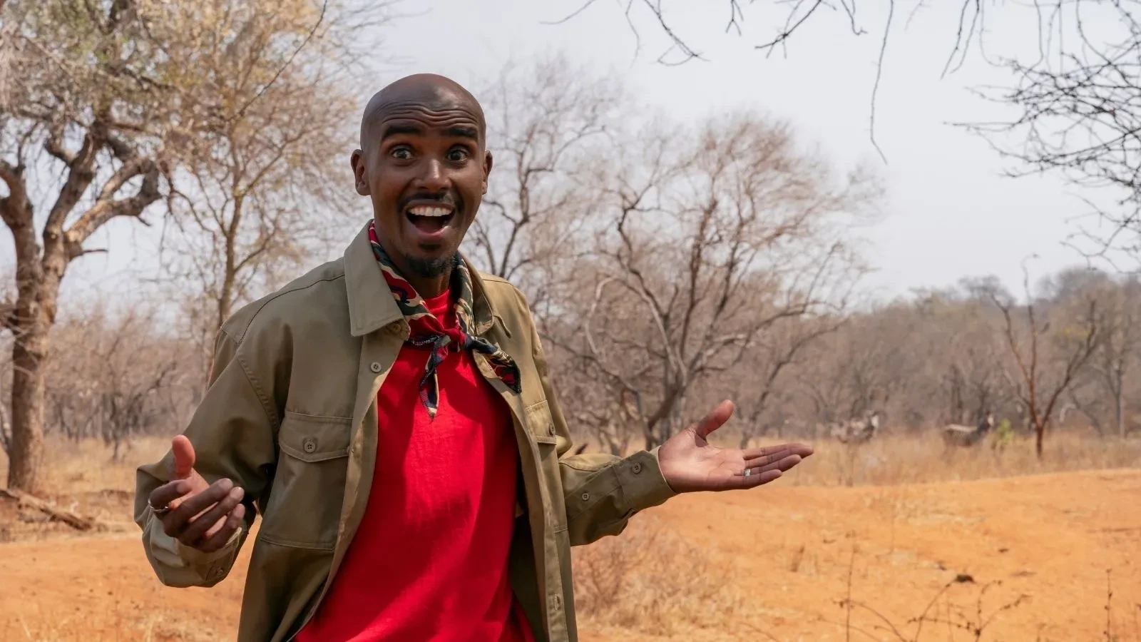 Sir Mo Farah standing in a dry, open landscape with trees behind him, wearing a red T‑shirt and khaki shirt while gesturing with his hands.