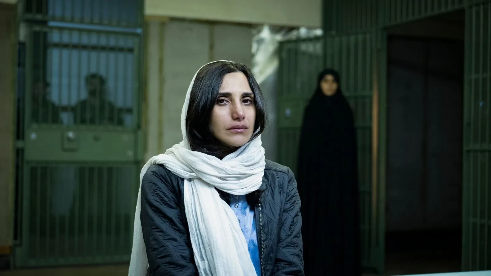 Individual wearing a light scarf seated inside a prison or detention facility with barred doors behind them.