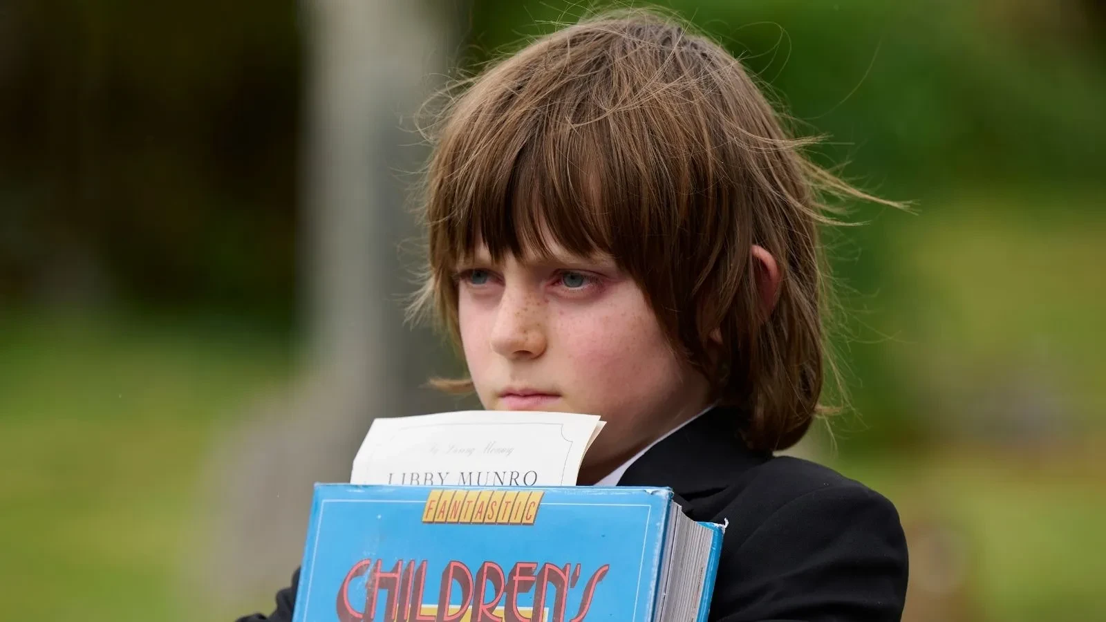 Child holding a large blue children's book outdoors, with long hair partially covering the face, standing against a green, natural background.