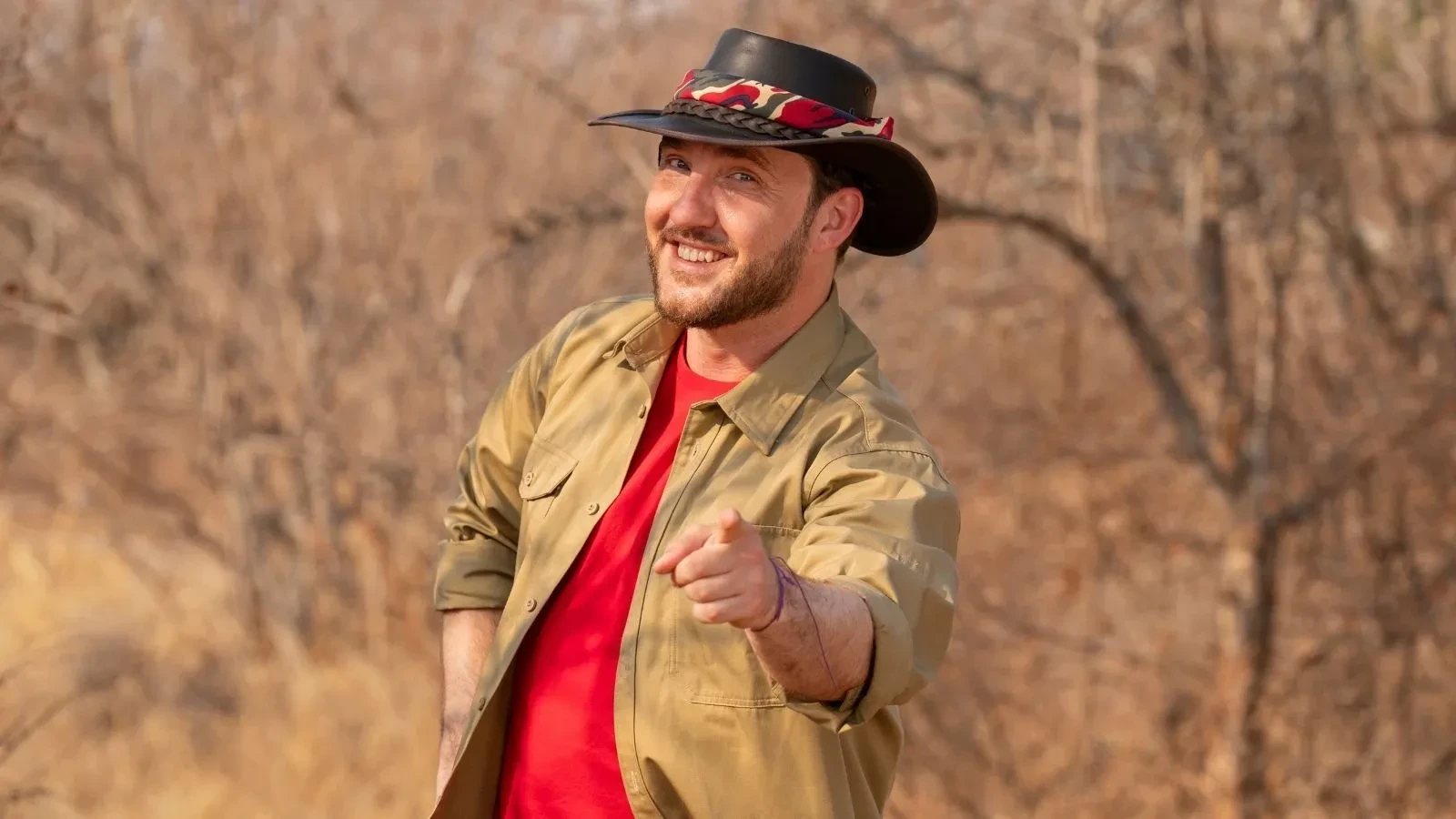 Seann Walsh pointing forward while standing in the South African bush, dressed in a red T‑shirt, khaki shirt, and wide‑brimmed hat.