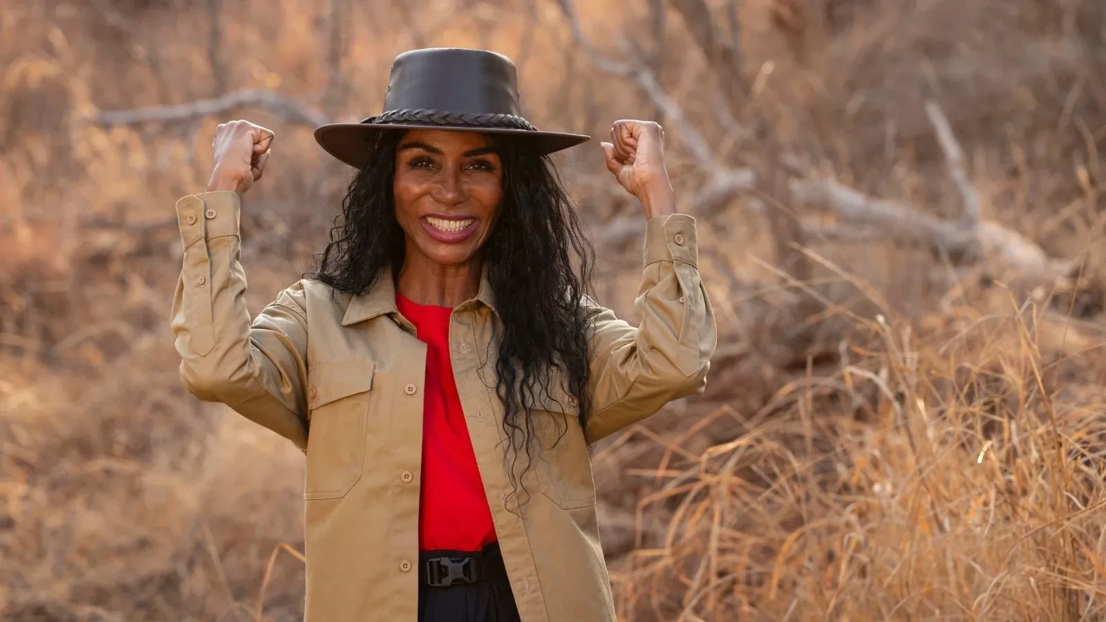 Sinitta posing confidently in the South African bush, wearing a wide‑brimmed hat, red top, and khaki shirt with arms raised in a strong gesture.