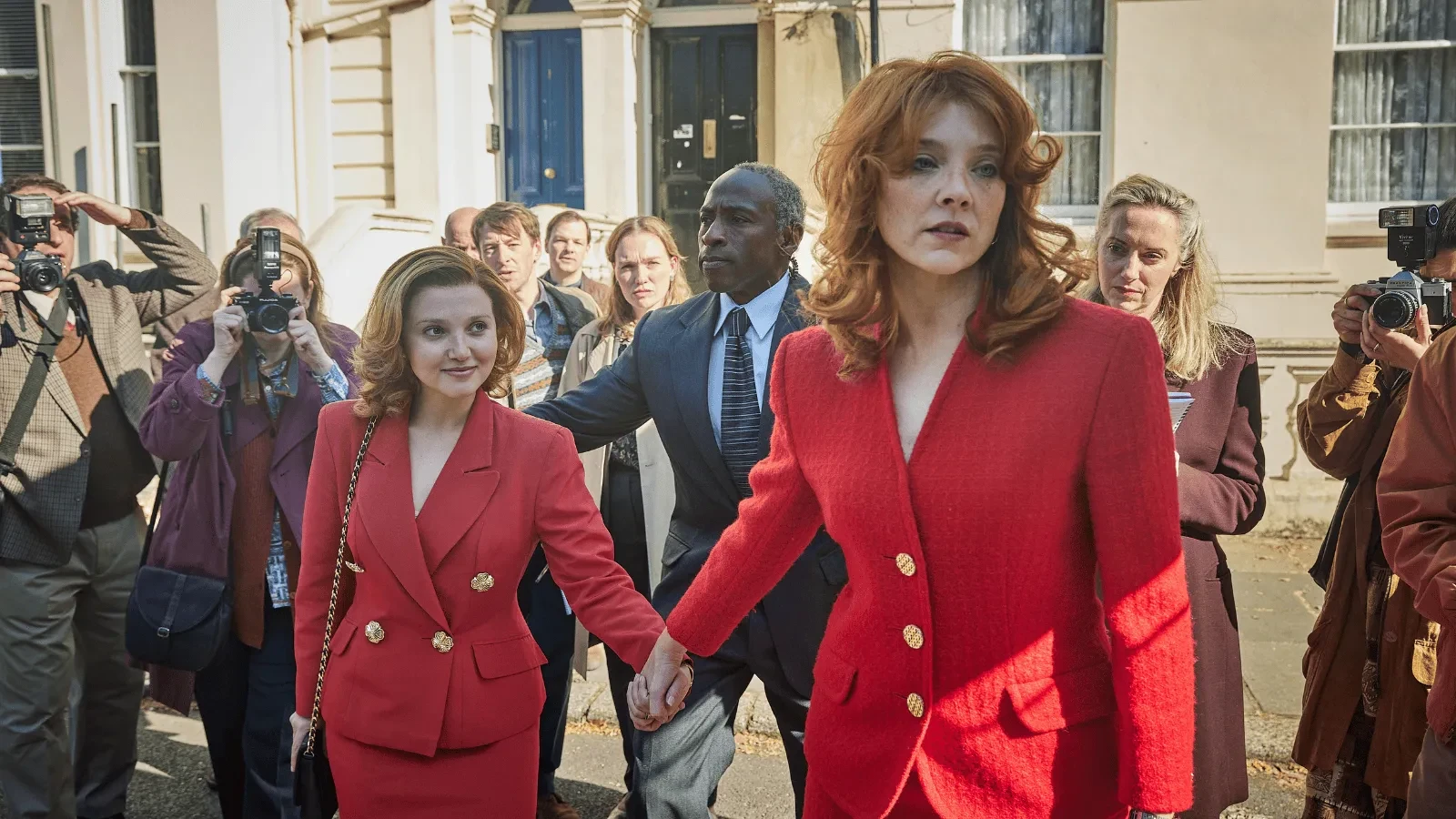 Two individuals in coordinated red outfits walk hand in hand down a street while surrounded by photographers capturing their entrance. 