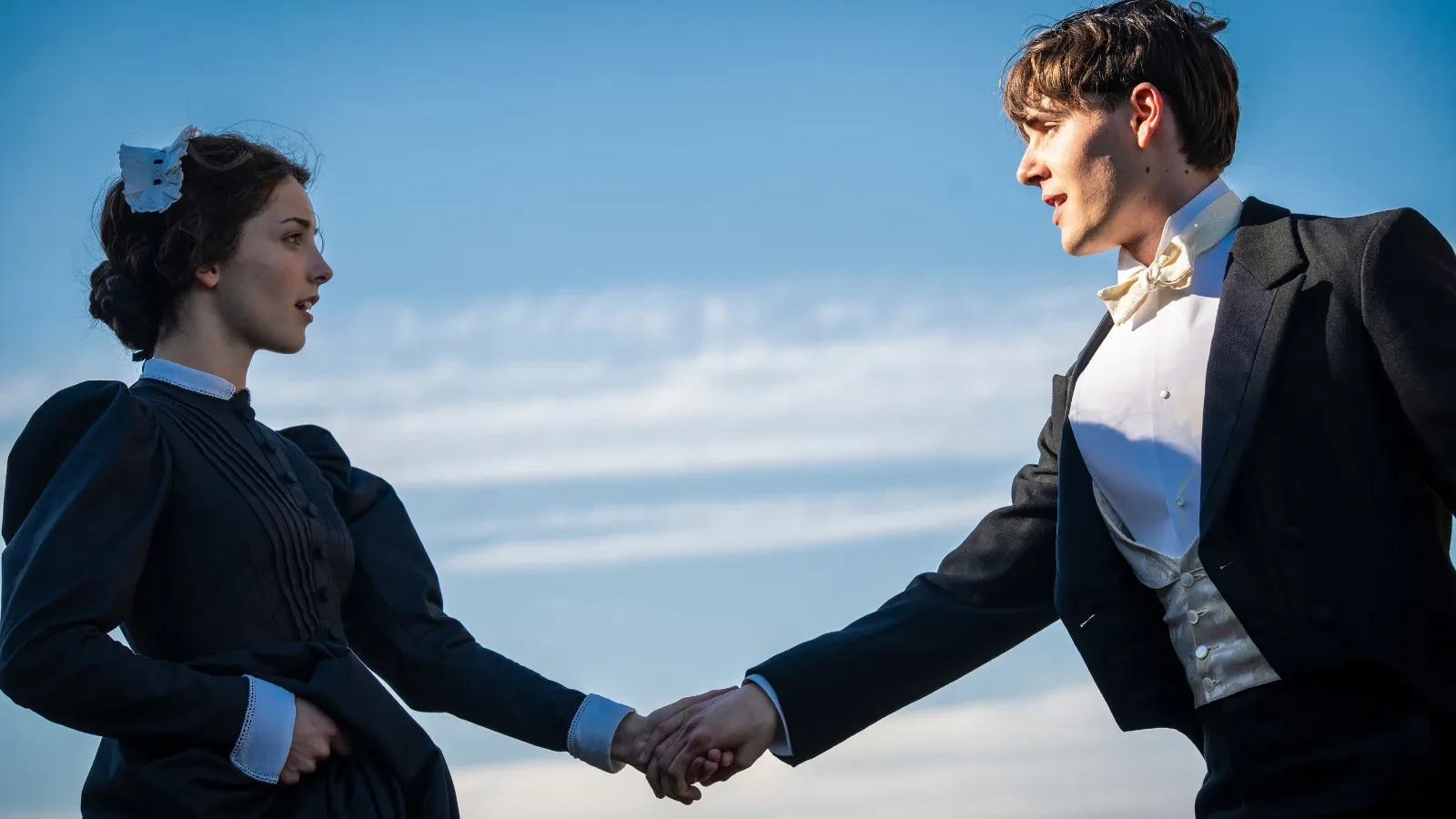 Two people in Victorian-style formal clothing stand outdoors, reaching out and holding hands against a blue sky with light clouds. 