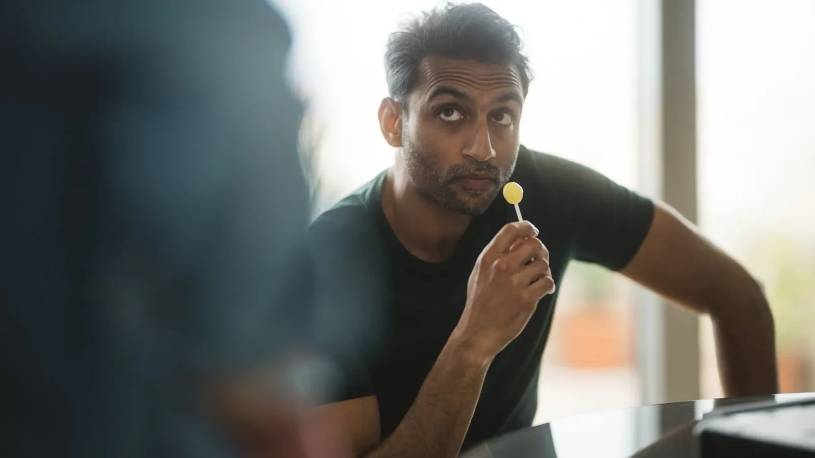Intelligence operative leaning on a table while holding a lollipop in a contemporary interior, scene from Secret Service TV series.