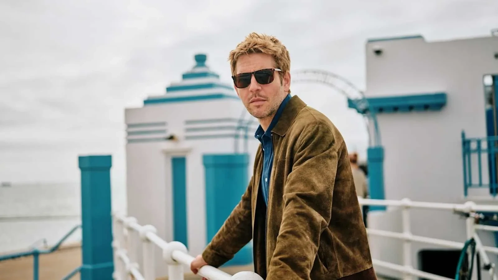 Jim Bergerac leans on railings at a seaside promenade, framed by Art Deco architecture in the Channel Islands drama Bergerac.
