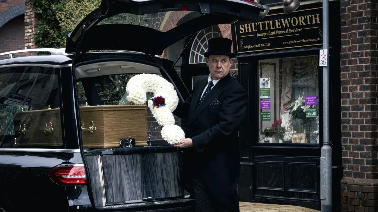 A hearse is parked outside a brick funeral home as a person lifts a white floral wreath beside a wooden coffin in the open rear of the vehicle. 