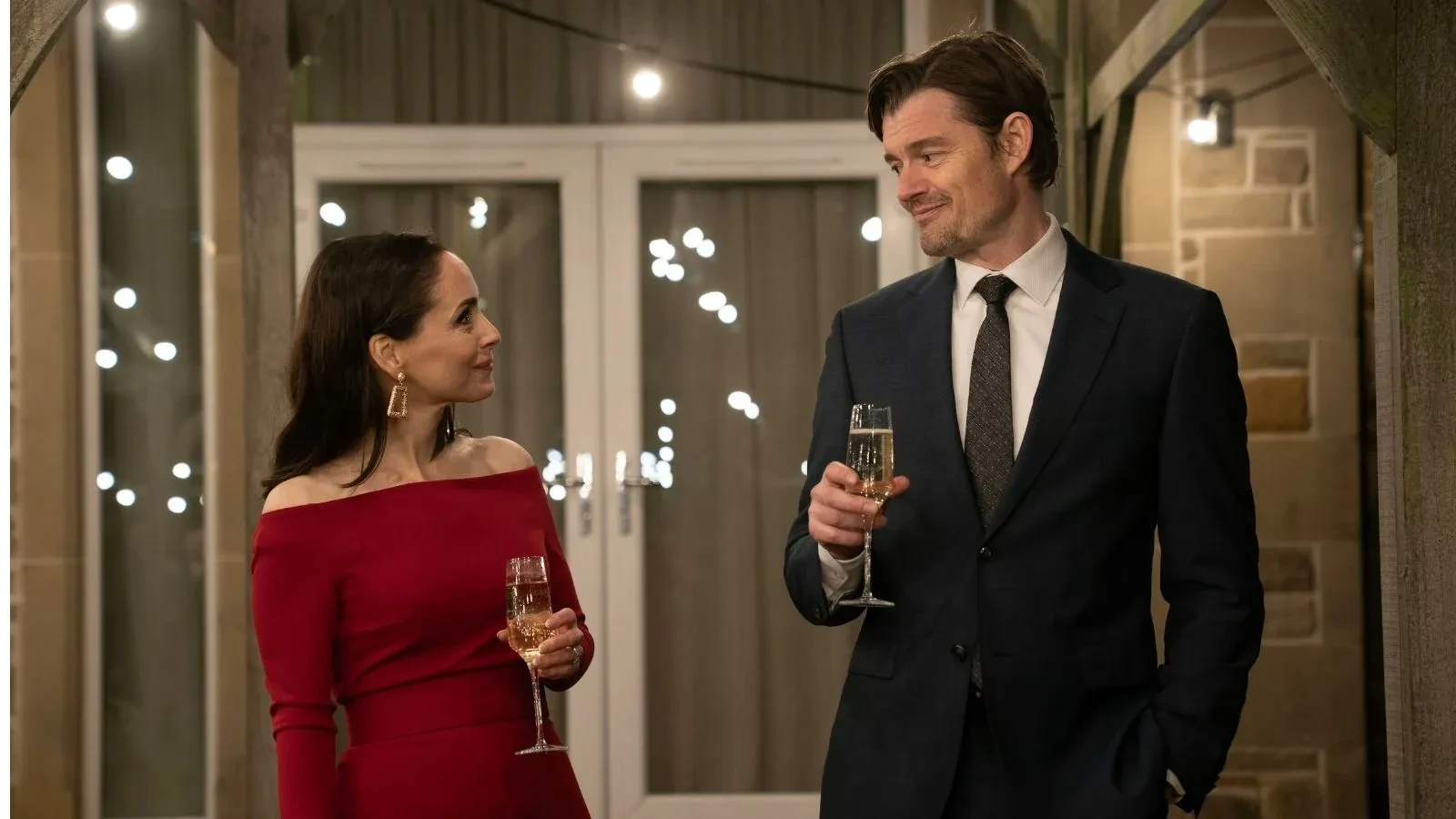 Two formally dressed people standing indoors at an evening event, each holding a glass of sparkling wine beneath warm string lighting.