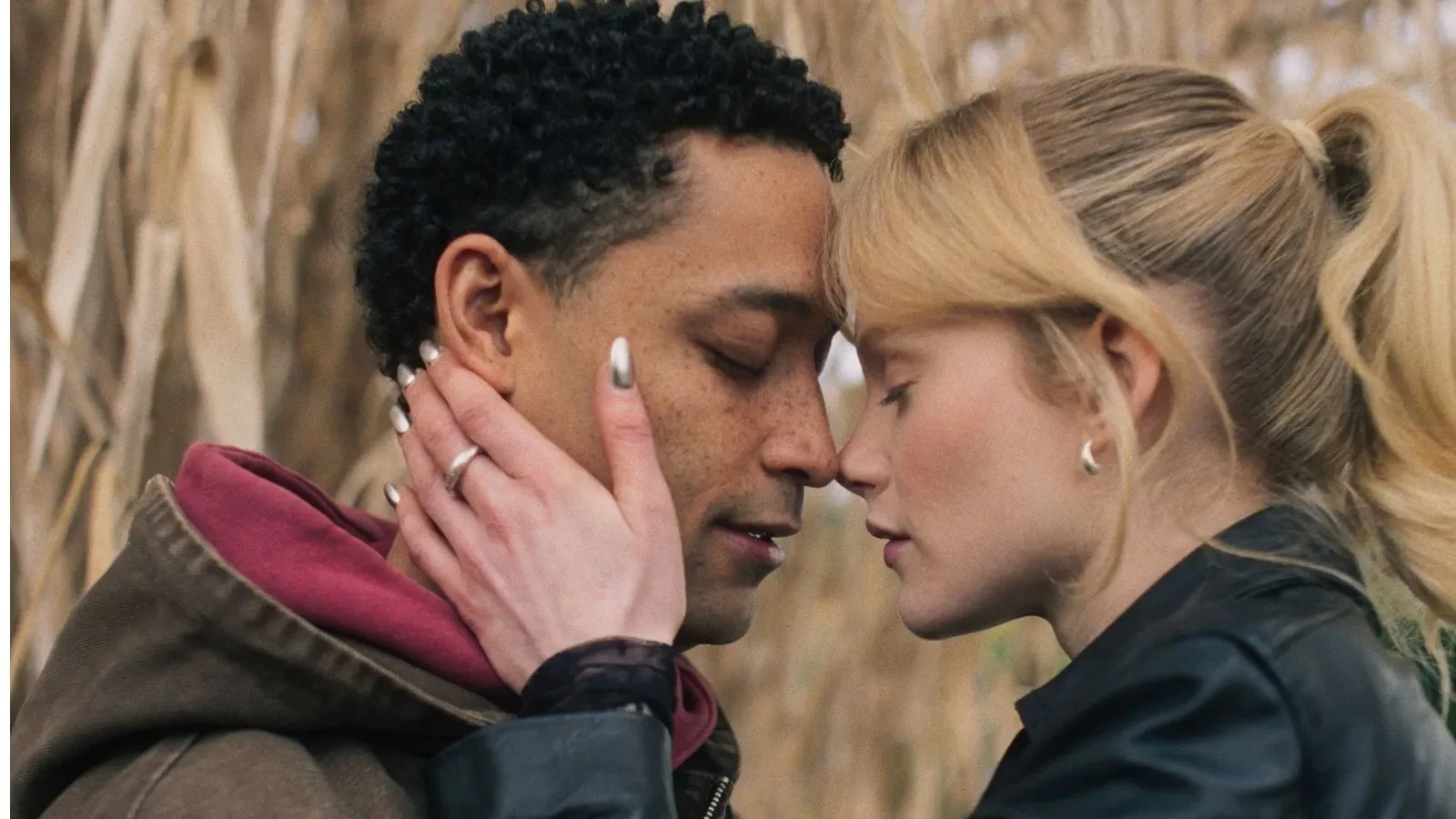 Close-up of two people standing outdoors among tall plants, touching foreheads, with one person’s hand gently placed on the other’s face.