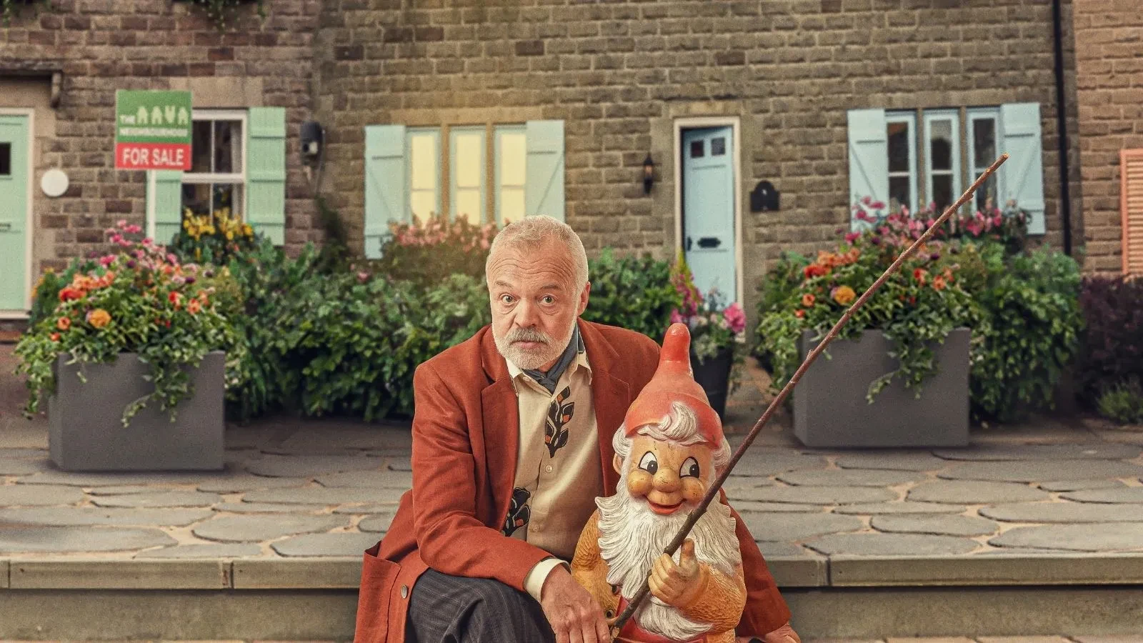 Graham Norton in a red jacket sat on steps holding a gnome with a fishing rod outside a nice looking house