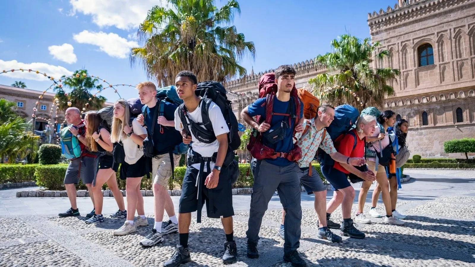 The contestants in Race Across the World season 6 wearing summer clothing abd backpacks posed ready to run by tropical trees and an old Italian style building