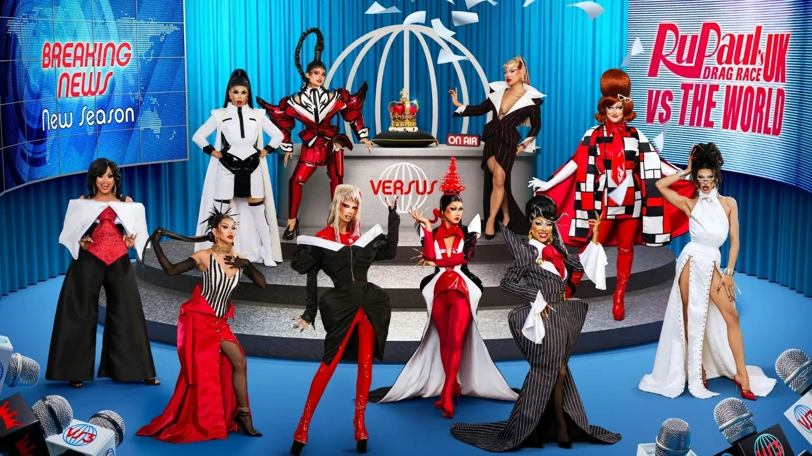 Drag queens in a newsroom dressed in extravagant black and white and red outfits with writing on screens on the walls that says "RuPaul's Drag Race UK vs The World"