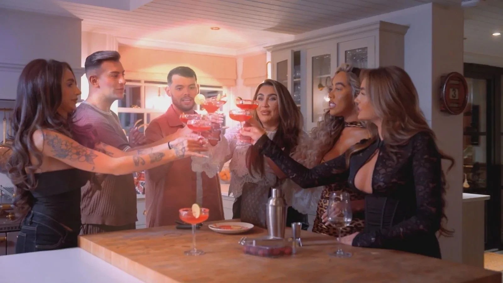 A group of people standing around a kitchen island indoors, raising pink cocktails in coupe glasses during a social gathering with warm lighting and neutral décor.