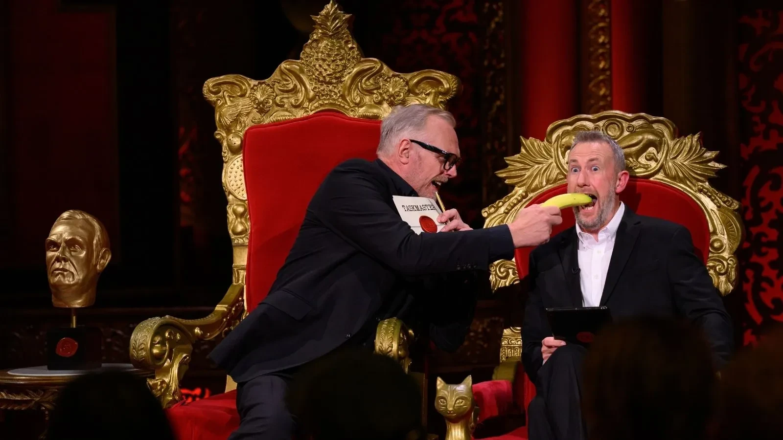 Two men sat on Taskmaster chairs with one holding a banana up to the mouth of the other man whilst laughing