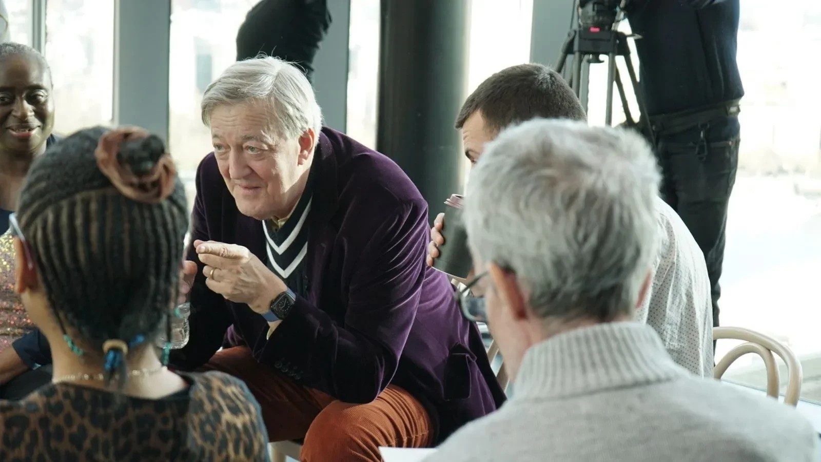 Stephen Fry wearing a purple velvet jacket and red corduroy trousers, sat on a chair with people gathering around him listening