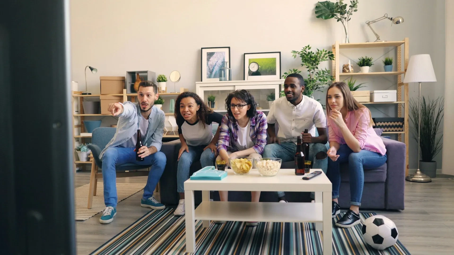 Five football fans sit on the sofa watching and reacting to a match on their TV