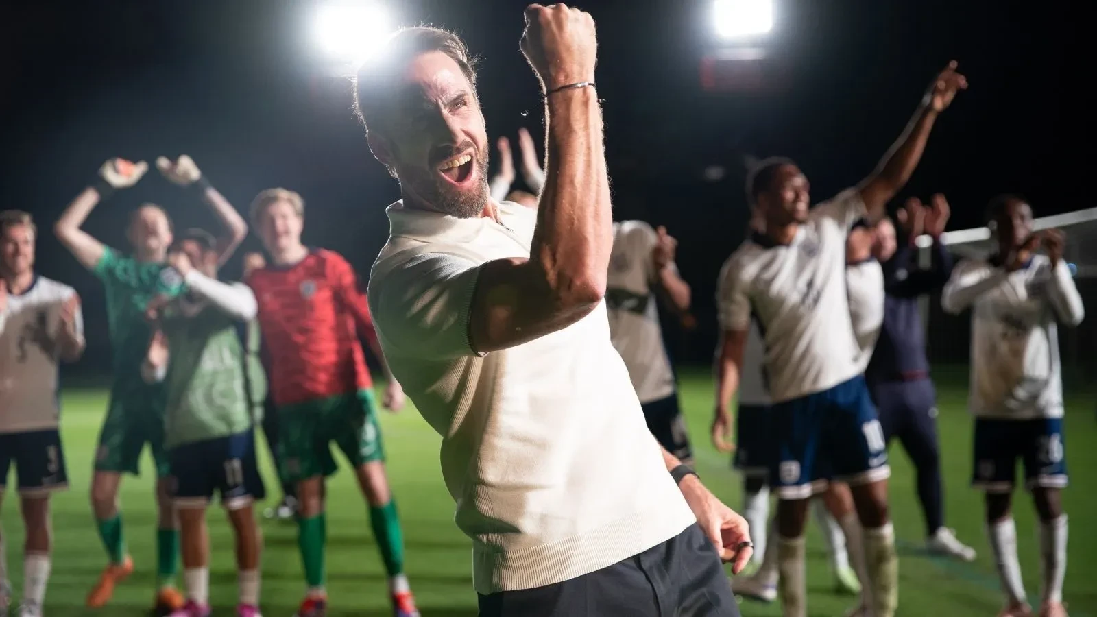 Promotional image for the drama Dear England showing a football coach celebrating on a floodlit pitch, with a team of players cheering in the background during a night match.