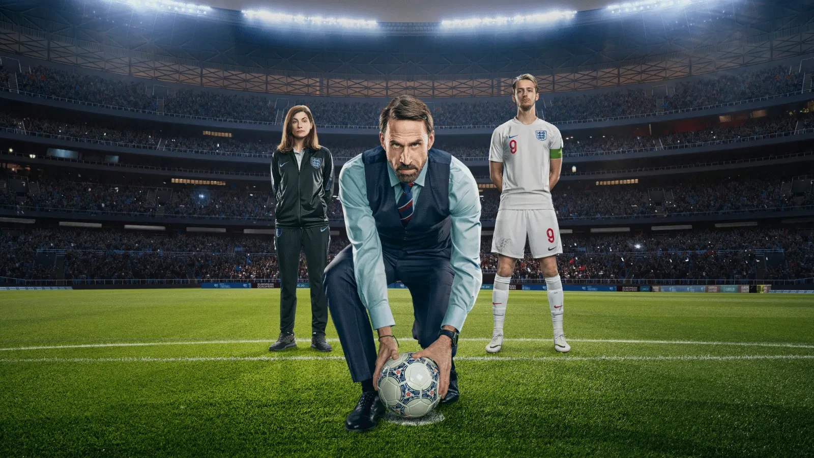 Joseph Fiennes stood in a smart outfit holding a football on a football pitch with a female coach behind him to the left and an England football player to the right