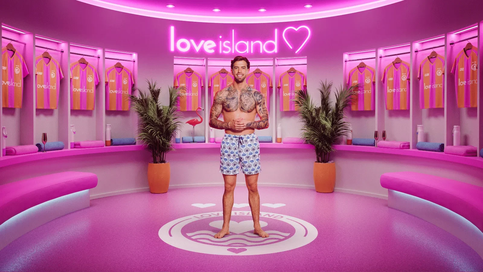 Aidan from season 13 of Love Island stood in the pink Love Island locker rooms with Love island Football shirts behind him