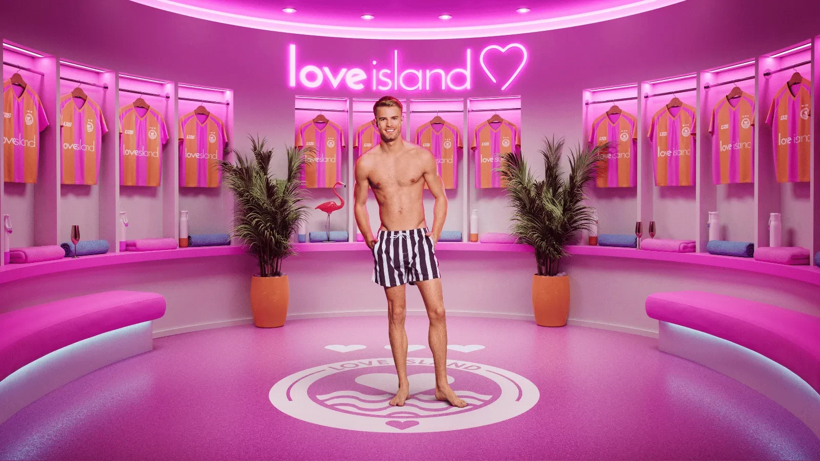 Sam from season 13 of Love Island stood in the pink Love Island locker rooms with Love island Football shirts behind him