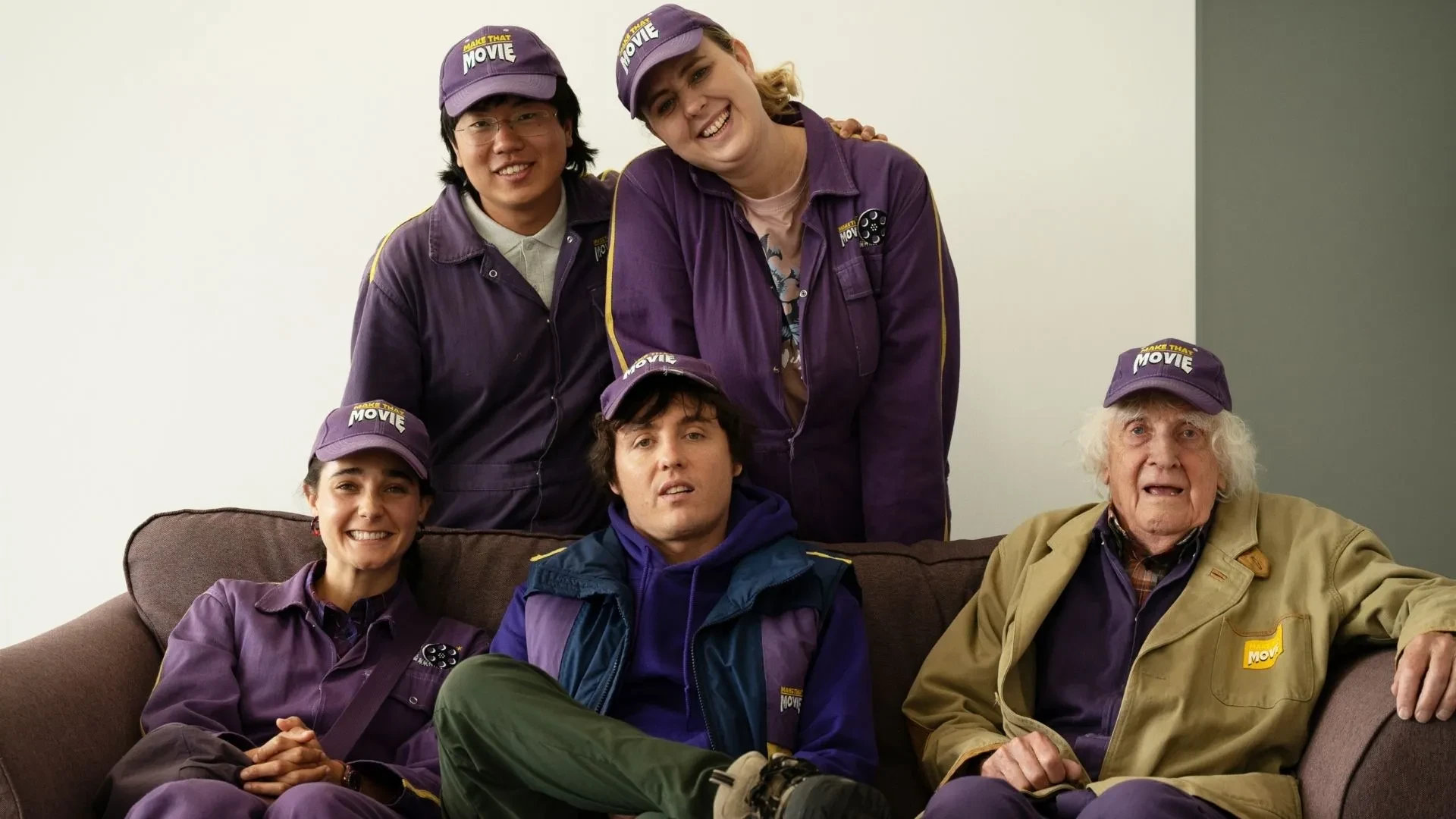 Ensemble cast image from Make That Movie showing six characters in matching purple uniforms sitting together indoors.