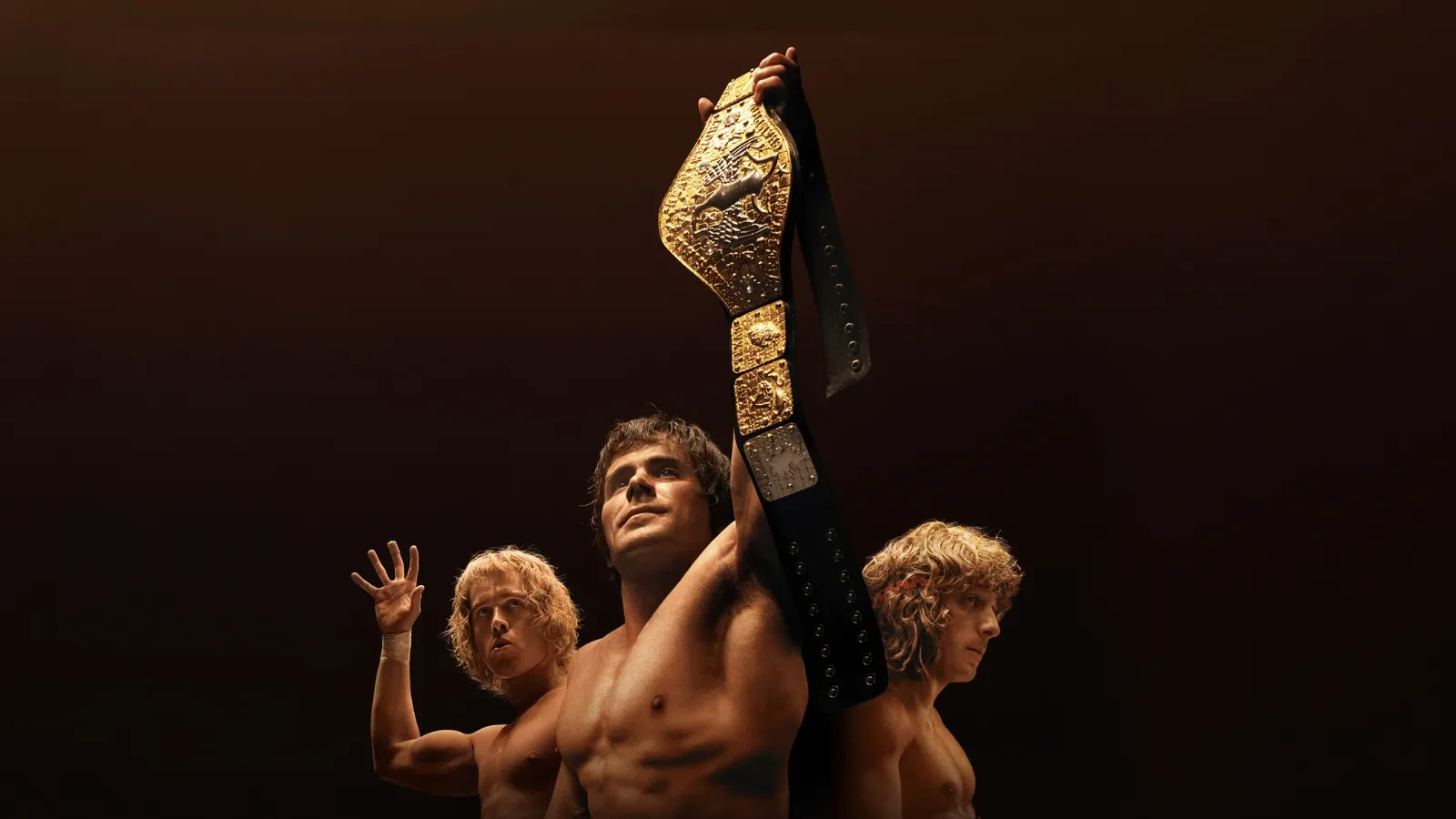 A man holding a golden wrestling belt with two young blonde men behind him against a dark background