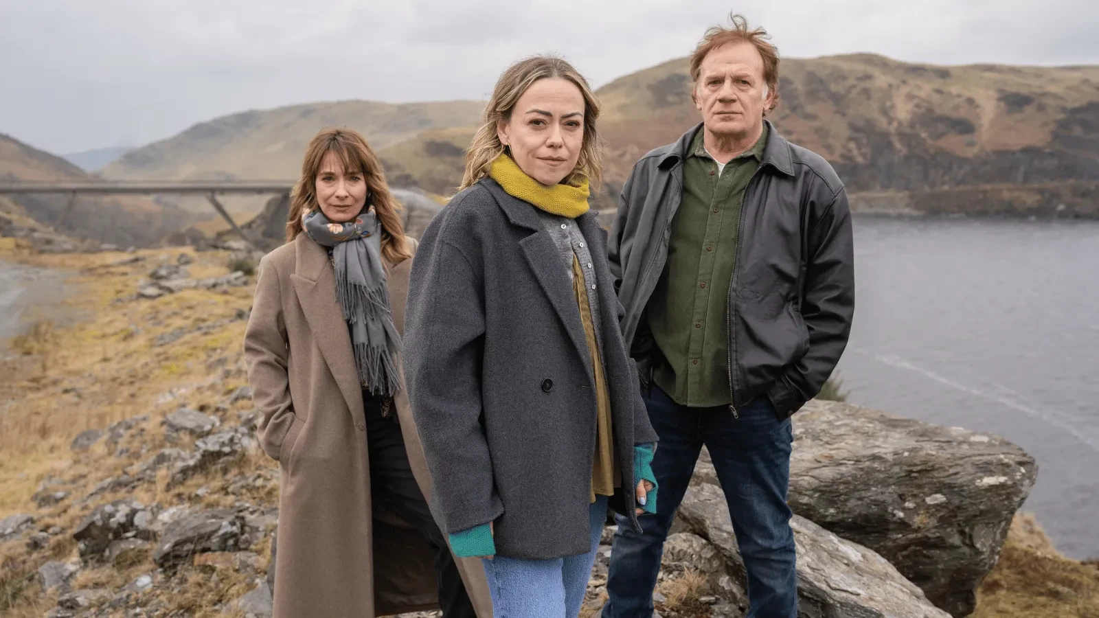 Two woman and a man in a warm clothing stood on a cliff top looking into the camera
