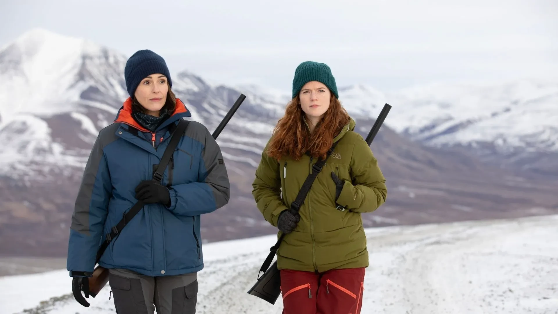 Crime thriller drama scene from Vigil showing two characters in cold-weather gear standing in a snowy mountain landscape.