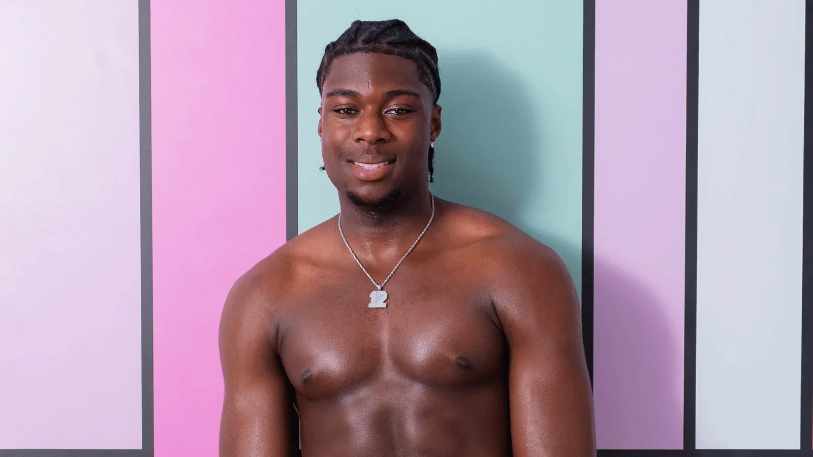 Smiling shirtless man with braided hair and a silver pendant necklace poses in front of a pastel geometric backdrop, facing the camera. 