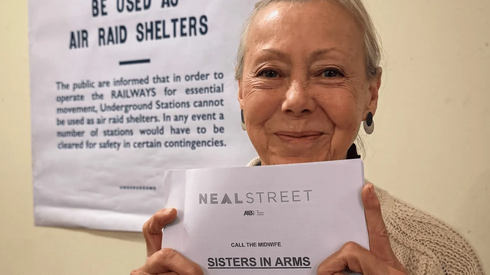 Person holding a Call the Midwife script titled Sisters in Arms, with a wartime air raid shelter poster visible in the background during a production-related event.